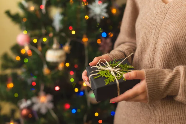 Mãos femininas segurando bom presente, árvore de Natal decorada no fundo — Fotografia de Stock