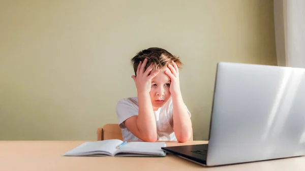 Child sits at desk, looks sadly at computer and holds his head with hands — Stock Photo, Image