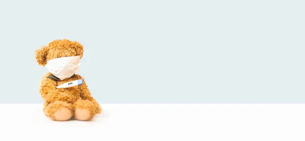 Urso de brinquedo senta-se em máscara no fundo de luz com termômetro — Fotografia de Stock