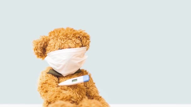 Panning vídeo de urso de brinquedo senta-se em máscara no fundo de luz com termômetro — Vídeo de Stock