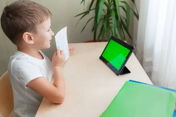 Anak sekolah pintar duduk di meja dan melihat tablet. Anak yang bahagia menunjukkan fotonya — Stok Foto