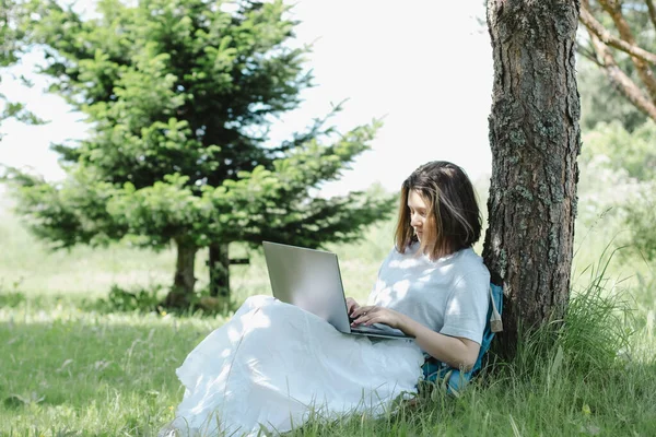 Dospívající dívka sedí v blízkosti stromu v letním parku s otevřeným notebookem v rukou — Stock fotografie