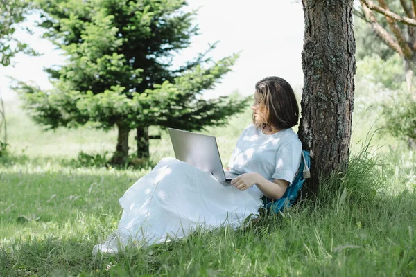 Dospívající dívka sedí v blízkosti stromu v letním parku s otevřeným notebookem v rukou — Stock fotografie