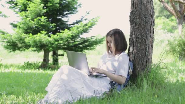 Ragazza adolescente si siede sull'erba vicino albero nel parco e stampe sul computer portatile. Giovane donna alza lo sguardo, sorride e agita la mano — Video Stock