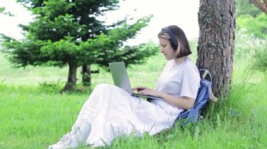 Genç kız dizüstü bilgisayarda yazıyor ve parkta ağaçların yanında otururken müzik dinliyor. Görüntüyü hafifçe büyüt