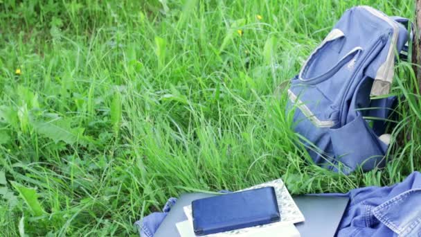 Mochila escolar, laptop, e-book, cadernos e camisa jeans deitado na grama verde. O vento balança a relva — Vídeo de Stock