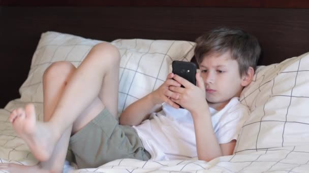 退屈少年はベッドに横たわって携帯電話で遊んで — ストック動画
