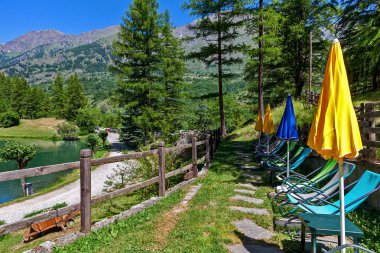 Kuzey İtalya, Piedmont 'taki yeşil ağaçların arasında küçük alp gölü boyunca sıralı sandalyeler ve şemsiyeler Laux.