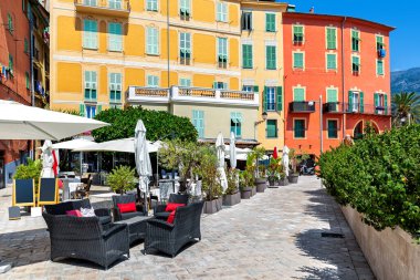 Fransa 'nın Menton şehrinde dar bir sokak, açık hava restoranı ve renkli evler görünüyor..