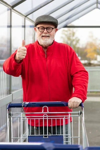 Uśmiechnięty Senior Czapką Czerwoną Kurtką Pchający Wózek Zakupy Podnoszący Kciuki — Zdjęcie stockowe