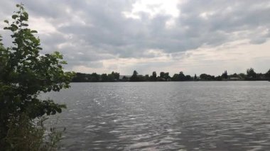 Sakin Trais Horloffer Gölü 'nün suları, bulutlu bir gökyüzü ve uzak bir köy kıyısının altında güneş yansımalarıyla dalgalanır.