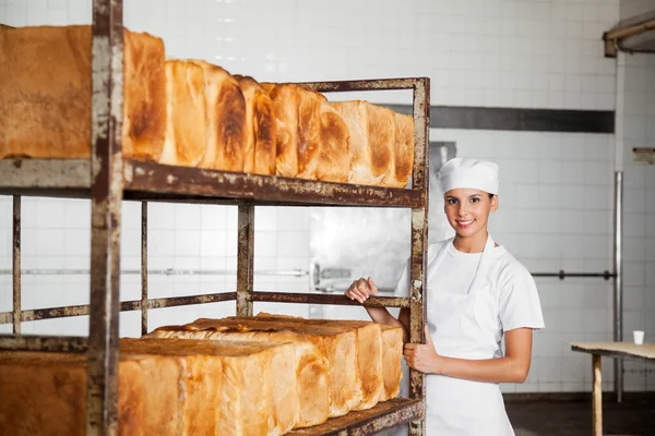 Panadero femenino de pie junto al bastidor con panes recién horneados — Foto de Stock