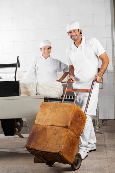 Trabajador empujando panes de pan en la carreta mientras su compañero de trabajo sonríe —  Fotos de Stock