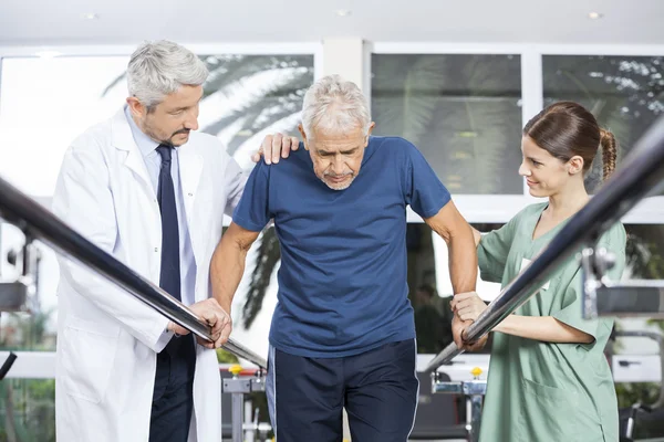 Fisioterapeutas motivando al paciente mayor a caminar entre parales —  Fotos de Stock