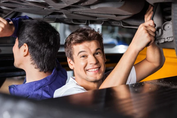 Mecánico feliz que trabaja debajo del coche levantado con colega —  Fotos de Stock