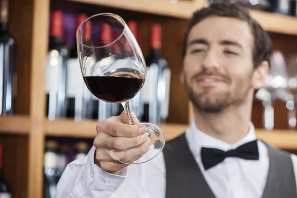 Jovem barman examinando vinho tinto em vidro — Fotografia de Stock