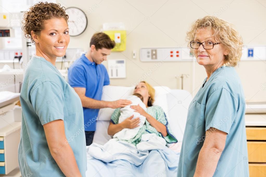 Successful Nurses Standing Against Couple With Baby In Backgroun