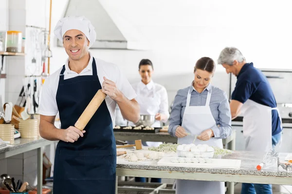 Chef masculino sonriente sosteniendo el rodillo mientras sus colegas se preparan — Foto de Stock