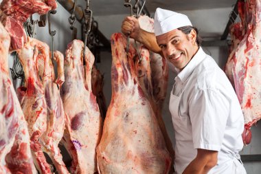 Butcher Standing By Meat Hanging In Slaughterhouse clipart