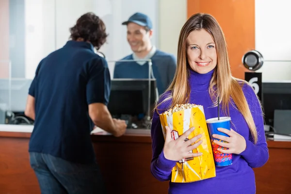 Mulher sorridente segurando lanches enquanto homem comprando ingressos de cinema — Fotografia de Stock