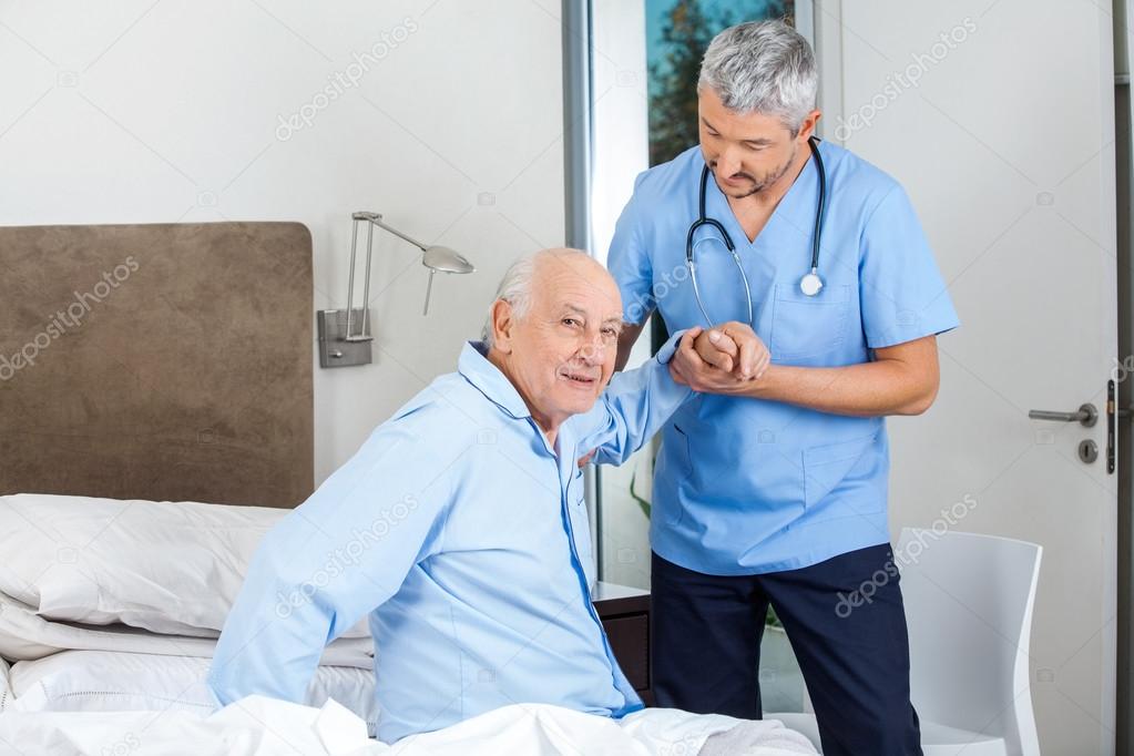 Senior Man Being Assisted By Male Caretaker In Bedroom