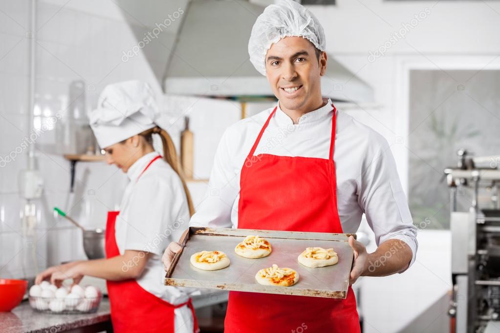 Smiling Chef Holding Small Pizzas On Baking Sheet At Kitchen