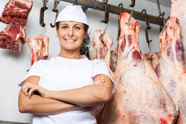 Carniceiro em pé contra a carne pendurada no açougue — Fotografia de Stock