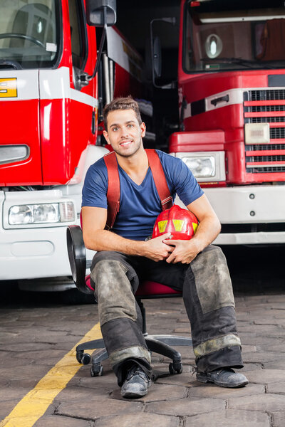 Smiling Firefighter Holding Helmet While Sitting Against Trucks