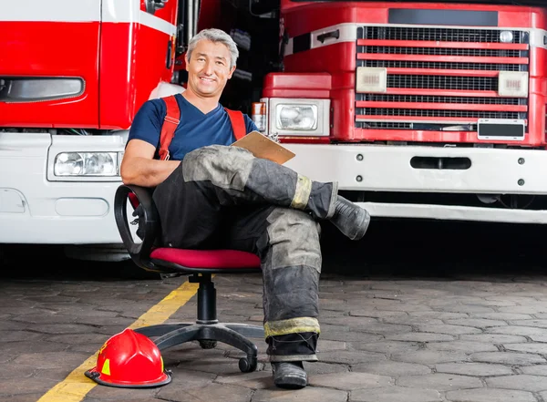 Happy Firefighter Sitting On Chair Against Trucks — Stock Photo, Image