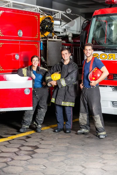 Jovens bombeiros felizes em pé contra caminhões — Fotografia de Stock