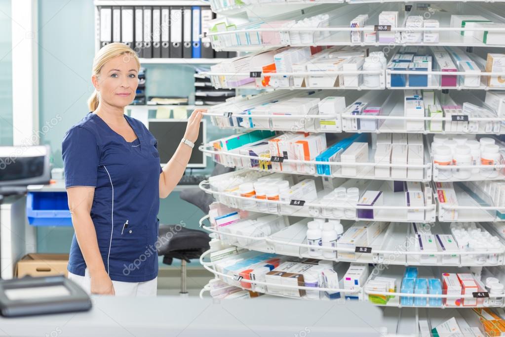 Female Chemist Standing By Shelves In Pharmacy
