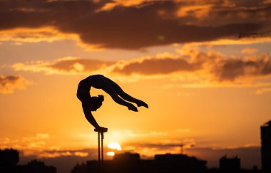 Esnek kadın sirk Artis dengeyi sağlar ve dramatik günbatımına ve şehir manzarasına karşı çatıda kıvrılır. Amuda kalkmak ve denge kavramı 