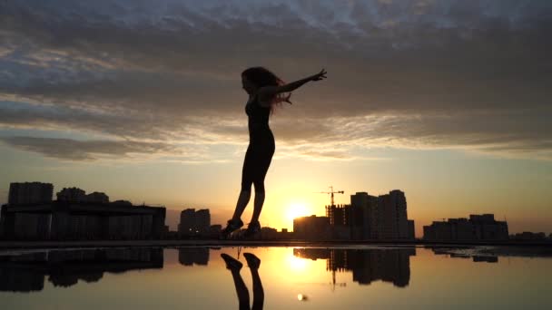 水面に映る街並みを背景に、夕暮れ時にゆっくりと動き回る女性の柔軟なダンサー。自由と幸福の概念 — ストック動画