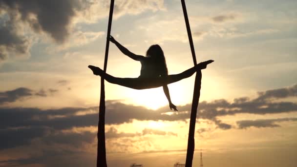 Silhouette of Female circus artist showing her flexibility and splits with red aerial silk on the dramatic sky background in slow motion. Concept of movement and motion — Stock Video