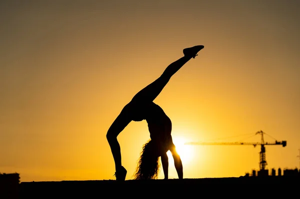 Gimnasta femenina mostrando su flexibilidad y dividida durante el atardecer sobre fondo de cielo naranja con grúa y construcción. Concepto de libertad y felicidad — Foto de Stock