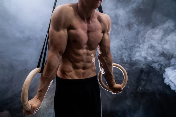 Muscular construir hombre haciendo crossfit en anillos de gimnasia interior en negro, fondo ahumado. concepto de motivación, deseo y pasión —  Fotos de Stock