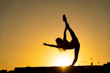 Kadın jimnastikçi esnekliğini gösteriyor ve turuncu arka planda gün batımında vinç ve inşaat ile ayrılıyor. Özgürlük ve mutluluk kavramı