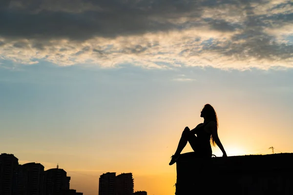 Silueta de mujer sentada en el borde de la azotea en el cielo dramático y el fondo del paisaje urbano durante la puesta del sol. Concepto de desafío y elección en la vida — Foto de Stock