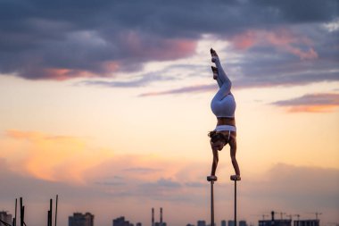 Esnek kadın sirk sanatçısı dramatik günbatımına ve şehir manzarasına karşı çatıda denge kuruyor ve kıvrılıyor. Amuda kalkmak ve denge kavramı 