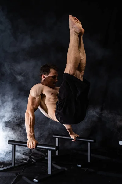 Muscular build man doing calisthenics on parallels bar indoor on black, smoked background. concept of healthy lifestyle and power