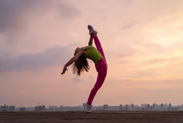 Mujer flexible haciendo split al aire libre en el dramático fondo de la puesta del sol. Concepto de yoga, estilo de vida saludable y estiramiento — Foto de Stock