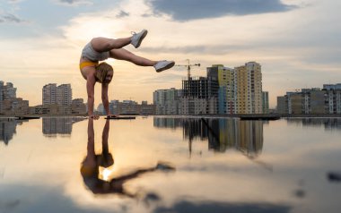 Esnek kadın jimnastikçi, dramatik günbatımında şehrin arka planında suda yansıma yaparak amuda kalkıyor ve jimnastik yapıyor.