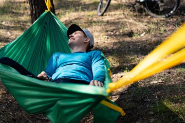 Genç, mutlu, şapkalı bir adam ormanda, hamakta dinleniyor..