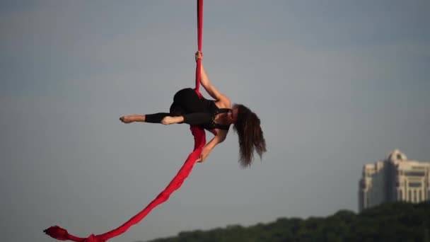 Цирковая артистка демонстрирует свою гибкость и расщепляется с красным воздушным шелком на фоне неба. Концепция здорового образа жизни — стоковое видео