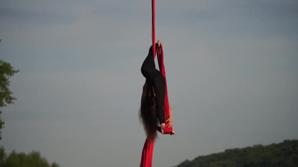 Женщина-акробат демонстрирует свою гибкость и расщепляется с красным воздушным шелком на фоне неба. Понятие грации, движения и красоты — стоковое видео
