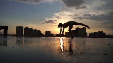 Esnek kadın jimnastikçi, akrobatik zıplayan, yağmurlu bir günde, şehir manzaralı dramatik günbatımında suda yansıyan yansımalarla numaralar yapan. Korkusuzluk, cesaret ve özgürlük kavramı 