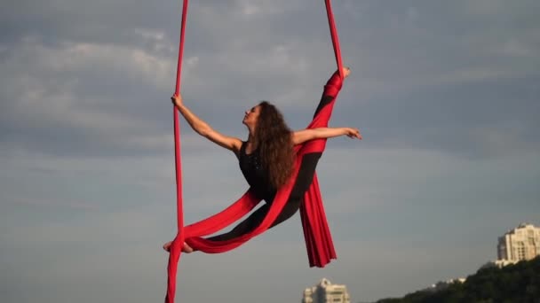 Kobieta akrobatka pokazuje swoją elastyczność i rozdziela się czerwonym jedwabiem lotniczym na tle nieba. Pojęcie łaski, ruchu i piękna — Wideo stockowe