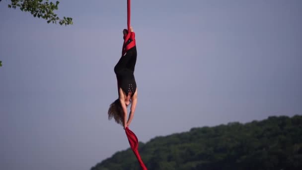 El acróbata femenino que muestra su flexibilidad y se divide con seda aérea roja en el fondo del cielo es de cámara lenta. Concepto de gracia, movimiento y belleza — Vídeo de stock