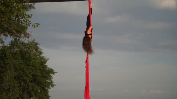 Fleksibel kvinnelig akrobat gjør et sprøtt og farlig triks med rød antenne silke. Solnedgangens himmelbakgrunn er langsom bevegelse. Vilje og lidenskap – stockvideo