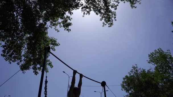 Silueta de hombre fuerte y musculoso haciendo trucos gimnásticos en barra horizontal en cámara lenta sobre el fondo azul del cielo. Concepto de estilo de vida saludable y calisténico — Vídeos de Stock